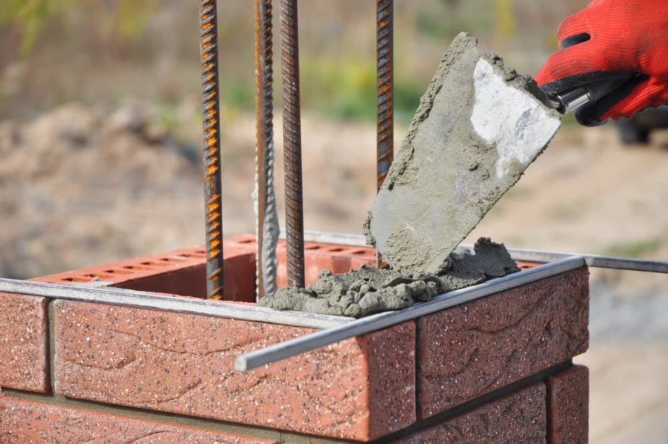 Chantier de construction en ext&eacute;rieur, ouvrier en gants rouges coulant du b&eacute;ton frais avec une truelle dans un moule d&eacute;limit&eacute; par des briques rouges perfor&eacute;es