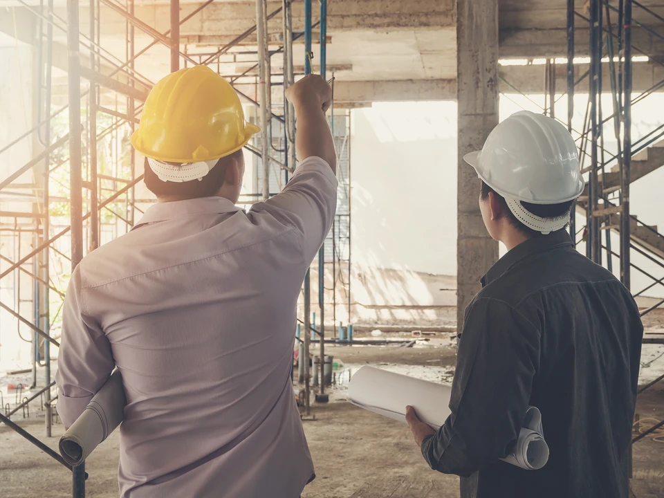Deux professionnels du bâtiment en casques de chantier jaune et blanc examinent un chantier de construction, l'un pointant vers une structure en béton armé
