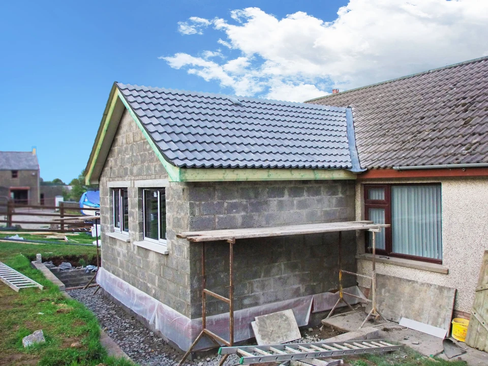 Extension de maison en construction avec murs en blocs de béton, toiture neuve en tuiles grises, échafaudage temporaire